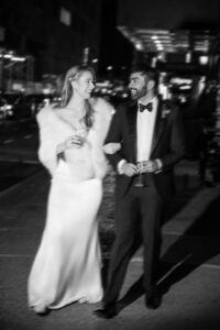 A couple in formal attire, holding drinks, walking together arm in arm on a city street at night. They are both looking at each other and smiling. The woman is wearing a long dress and fur stole; the man is wearing a tuxedo.