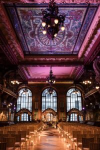 An elegant hall with a stained glass ceiling, chandeliers, and rows of chairs facing a floral arrangement. On the floor there are also candles.