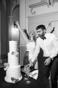 a couple at their wedding reception in front of them is their wedding cake the woman is wearing a white wedding dress and has both hands in the air the man is reaching towards the cake