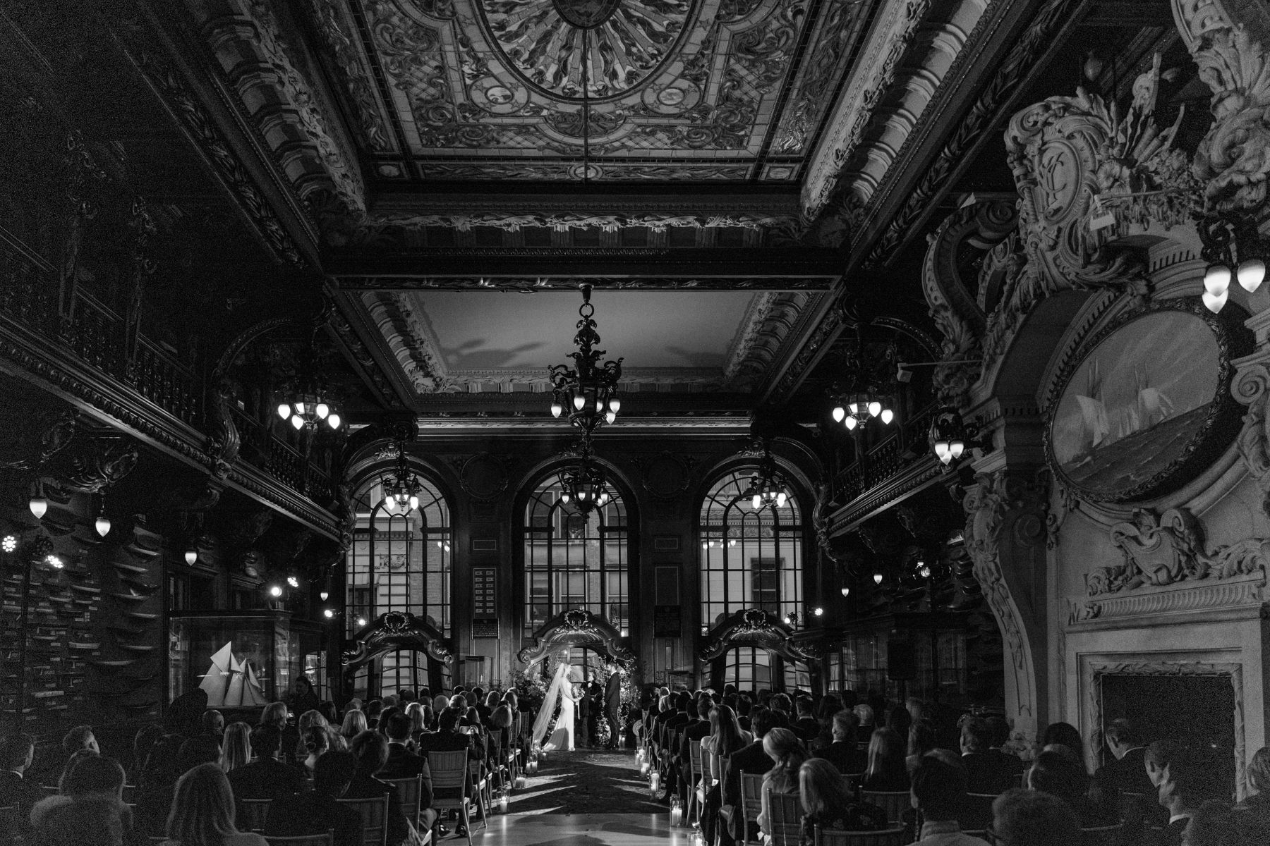 an elegant hall with large windows where a couple is having their wedding ceremony their wedding guests are seated in front of them and the couple is standing next to the bay window having their ceremony