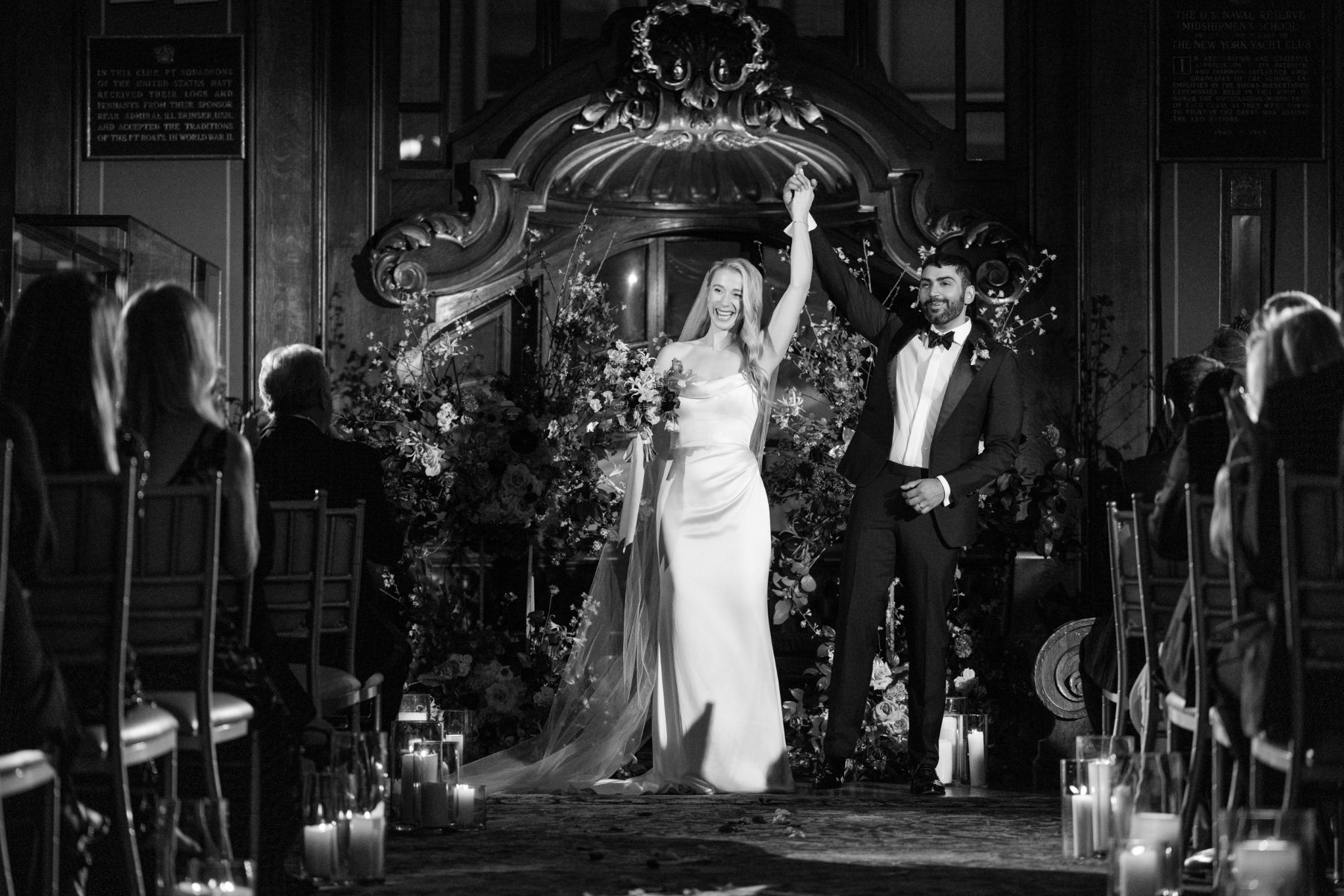 a couple at their wedding recepiton they are both holding their hands in the air the woman is wearing a white wedding dress and holding a bouquet of flowers the man is wearing a suit 