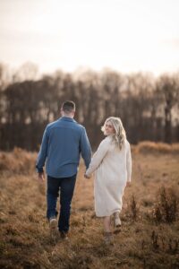 A couple strolls hand in hand through the grassy field, with trees in the background.