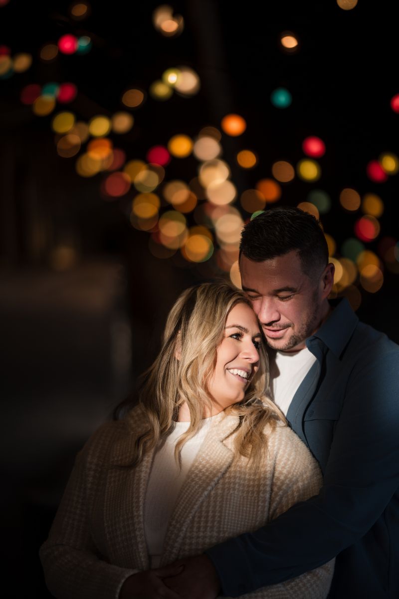 a couple standing together and smiling underneath twinkling lights the woman is leaning back into her partner