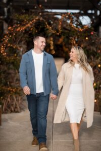 A couple strolls hand in hand, smiling at each other beneath a canopy of twinkling lights and greenery