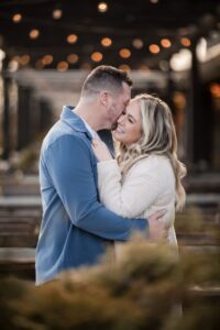 a couple standing together in a cafe the man is kissing the woman on the cheek and the woman is smiling behind them are twinkling lights