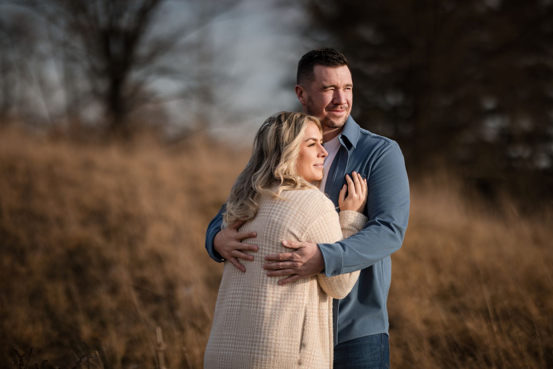 a couple standing together in a field hugging one another and looking off to the distance 