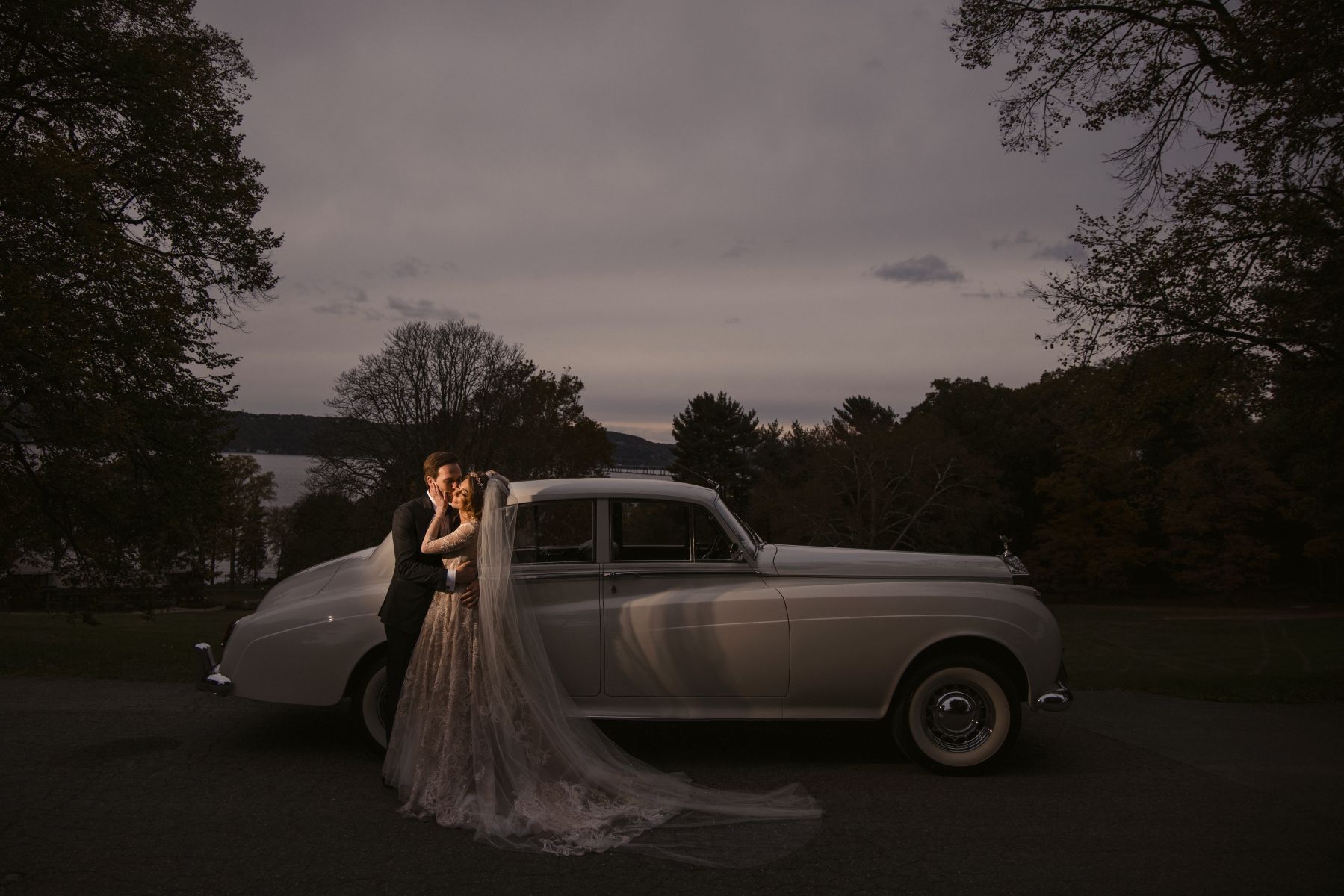 A couple embraces in front of a classic white car, the bride's is wearing a long veil draped over the ground.