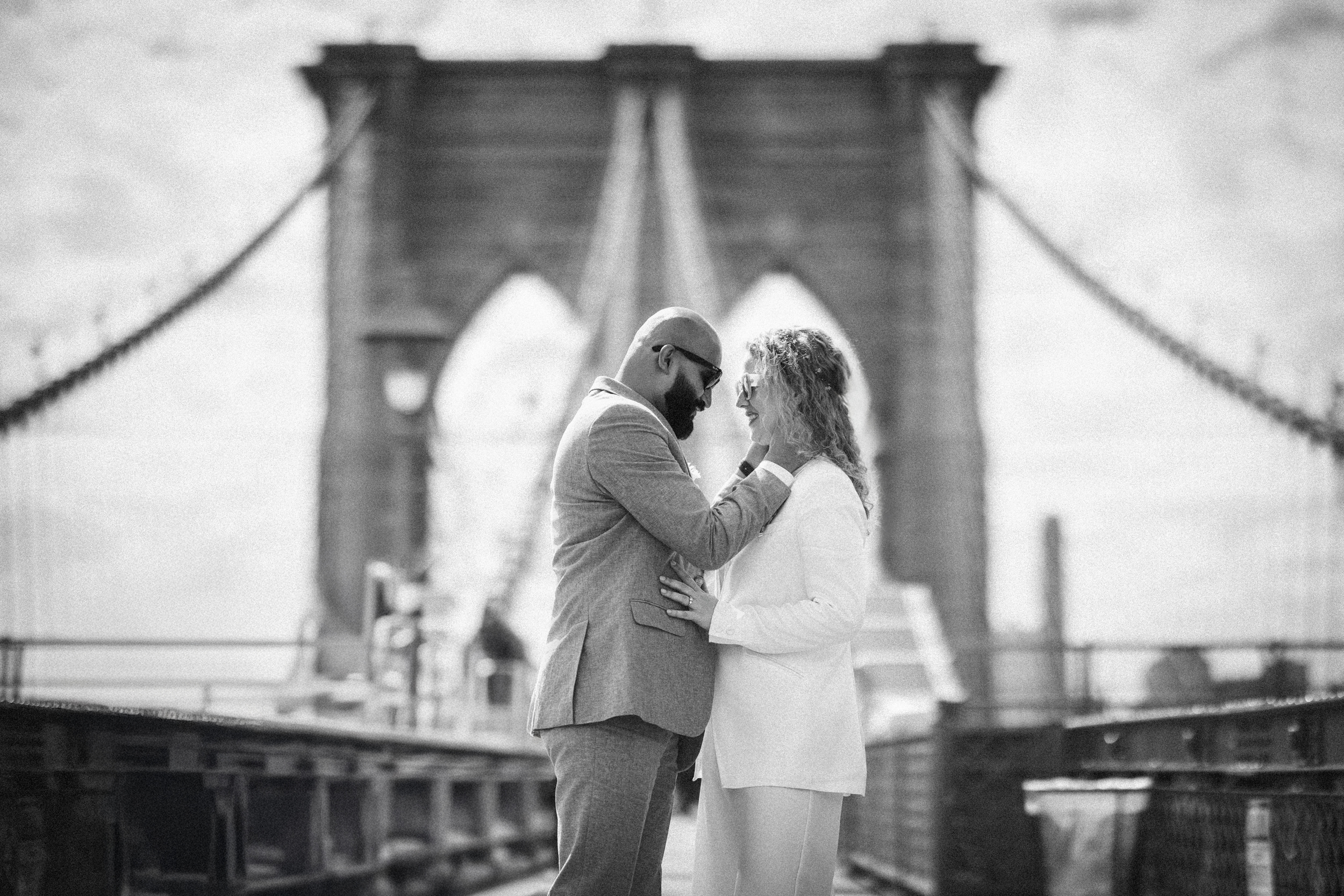 A couple stands facing each other on the Brooklyn Bridge in this black and white elopement photo, with one person gently touching the other's face.