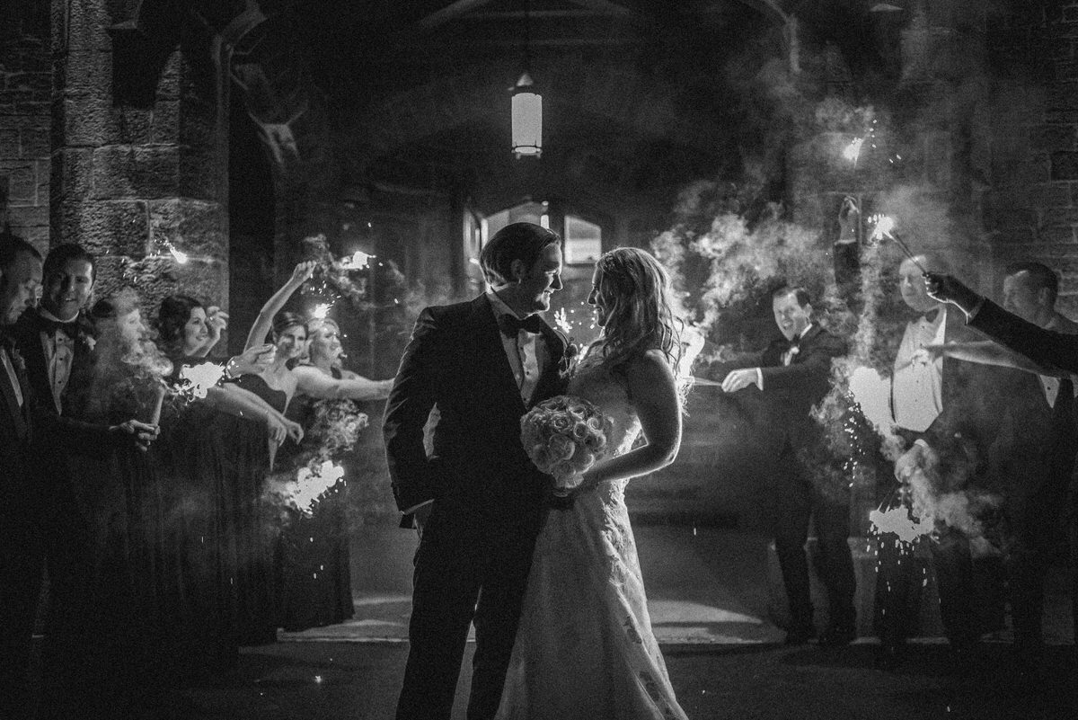 Newlywed couple with their arms around each other as their guests celebrate around them 