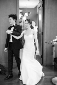 A couple in formal attire, with the woman in a strapless wedding dress and the man in a tuxedo, hold hands and smile as they make an entrace to their wedding reception