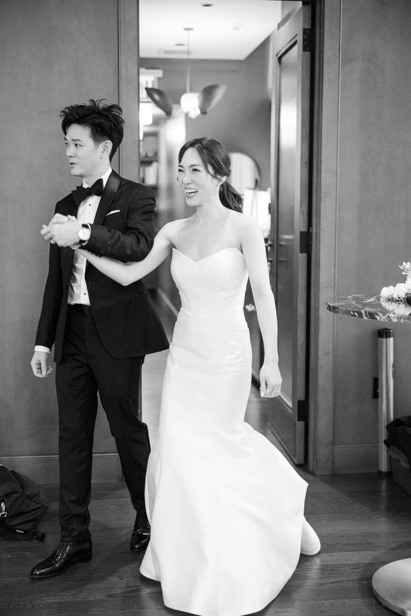 A couple in formal attire, with the woman in a strapless wedding dress and the man in a tuxedo, hold hands and smile as they make an entrace to their wedding reception