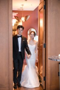 A couple dressed in formal wedding attire standing hand in hand in a doorway, ready to celebrate in the private dining room at ci siamo in new york city