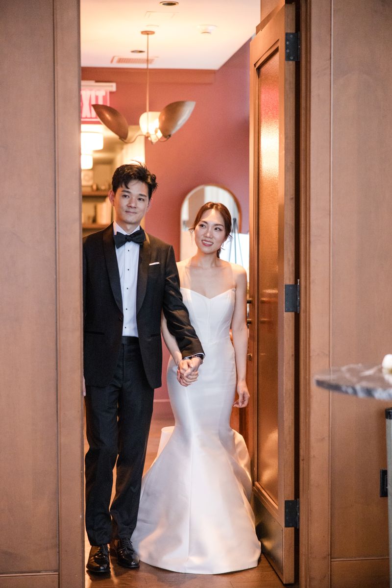 A couple dressed in formal wedding attire standing hand in hand in a doorway, ready to celebrate in the private dining room at ci siamo in new york city