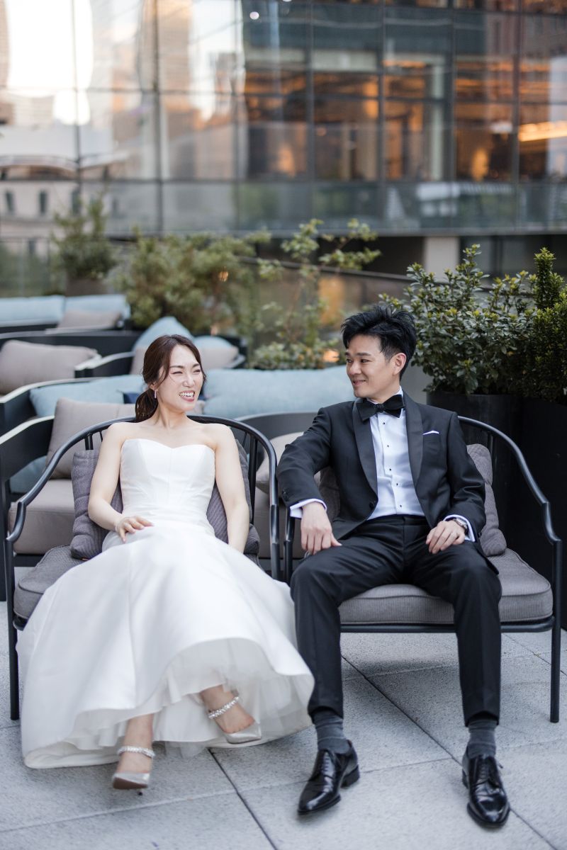 A bride in a white dress and a groom in a black tuxedo sit next to each other on outdoor lounge chairs, smiling and looking at each other at ci siamo in new york city