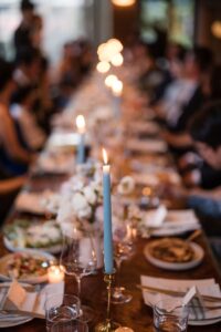 A long dining table set with plates, glasses, floral centerpieces, and lit candles, with people seated on both sides in a dimly lit setting