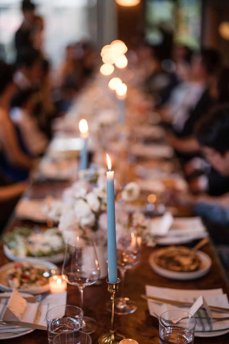 A long dining table set with plates, glasses, floral centerpieces, and lit candles, with people seated on both sides in a dimly lit setting