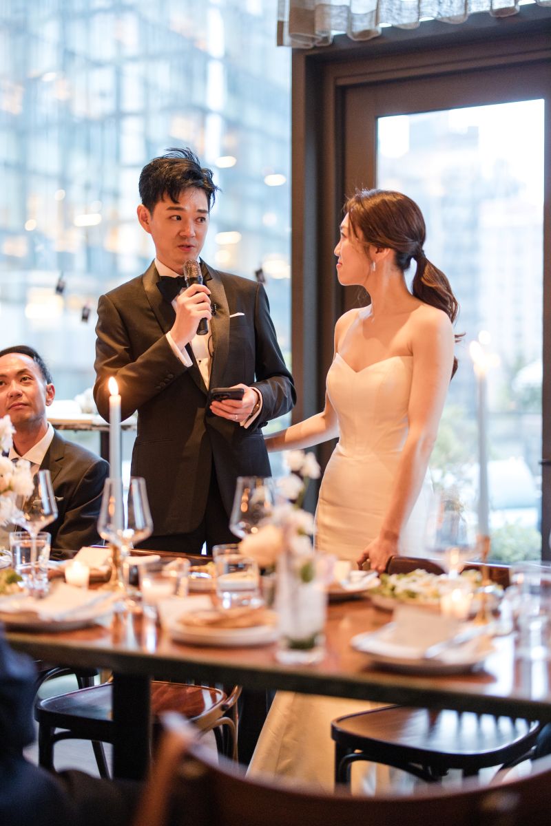 A man in a suit holds a microphone and speaks while standing next to a woman in a white wedding dress at an elegant dinner table