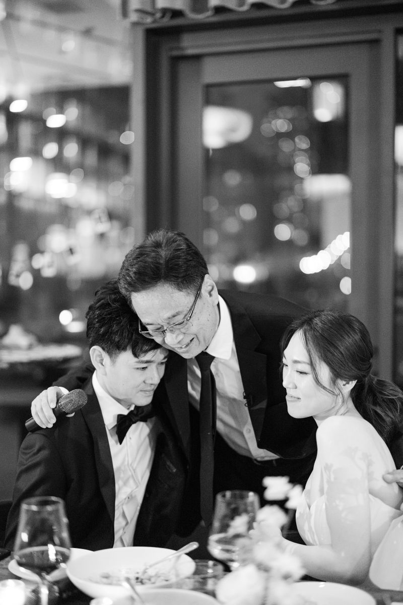 A man in a suit embraces a seated couple at their wedding reception