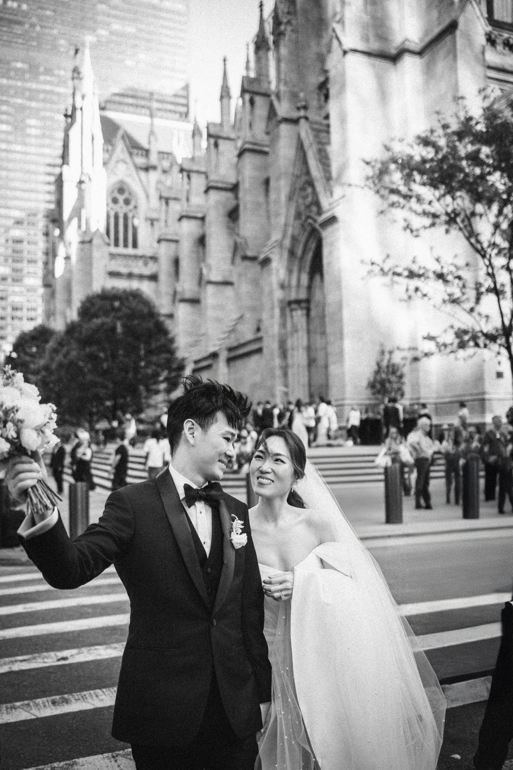 A bride and groom in wedding attire stand together on a city street in front of the iconic St. Patrick’s Cathedral