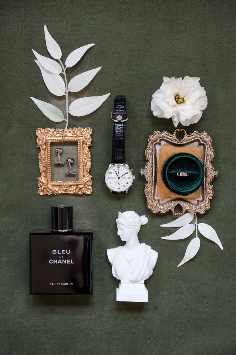 Flat lay of a watch, perfume, framed keys and ring, a white flower, green ring box, white leaves, and a white bust on a dark green background