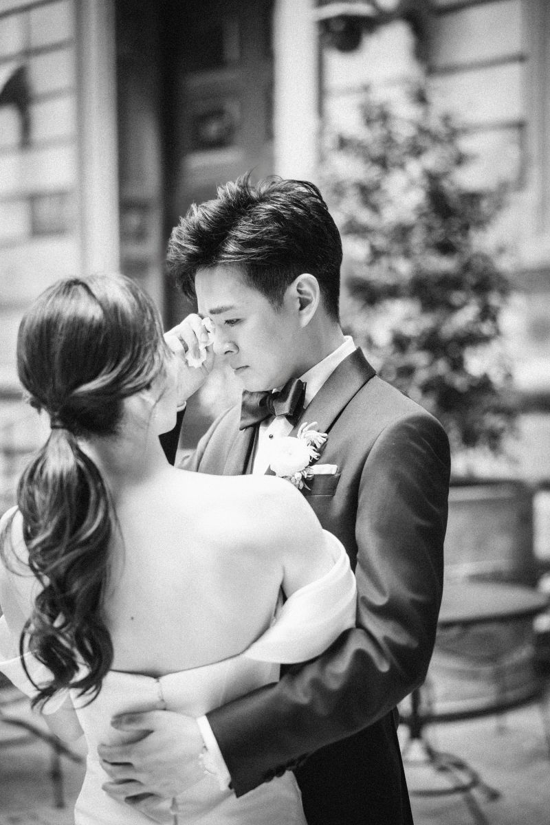 A bride and groom embrace outdoors at their wedding venue, with the bride gently wiping a tear from the groom’s face.