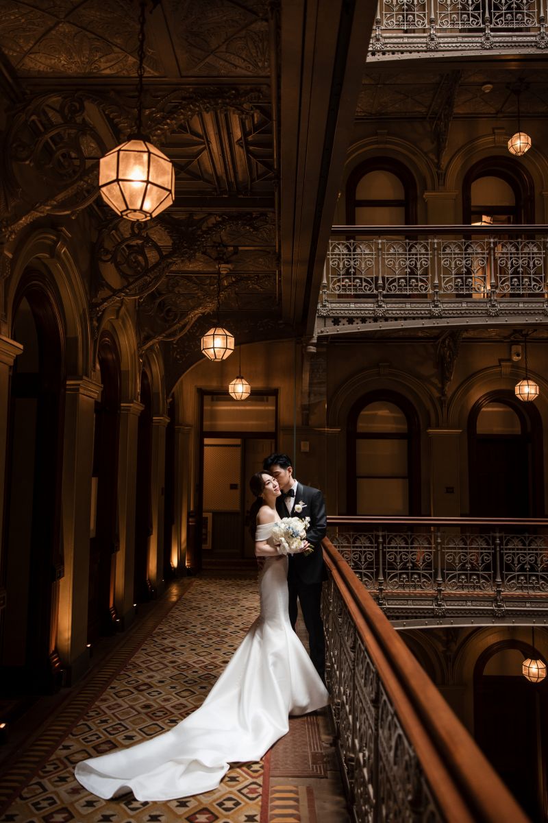 A bride and groom in formal attire embrace on an ornate balcony at a historic wedding venue, with decorative lighting and intricate architectural details