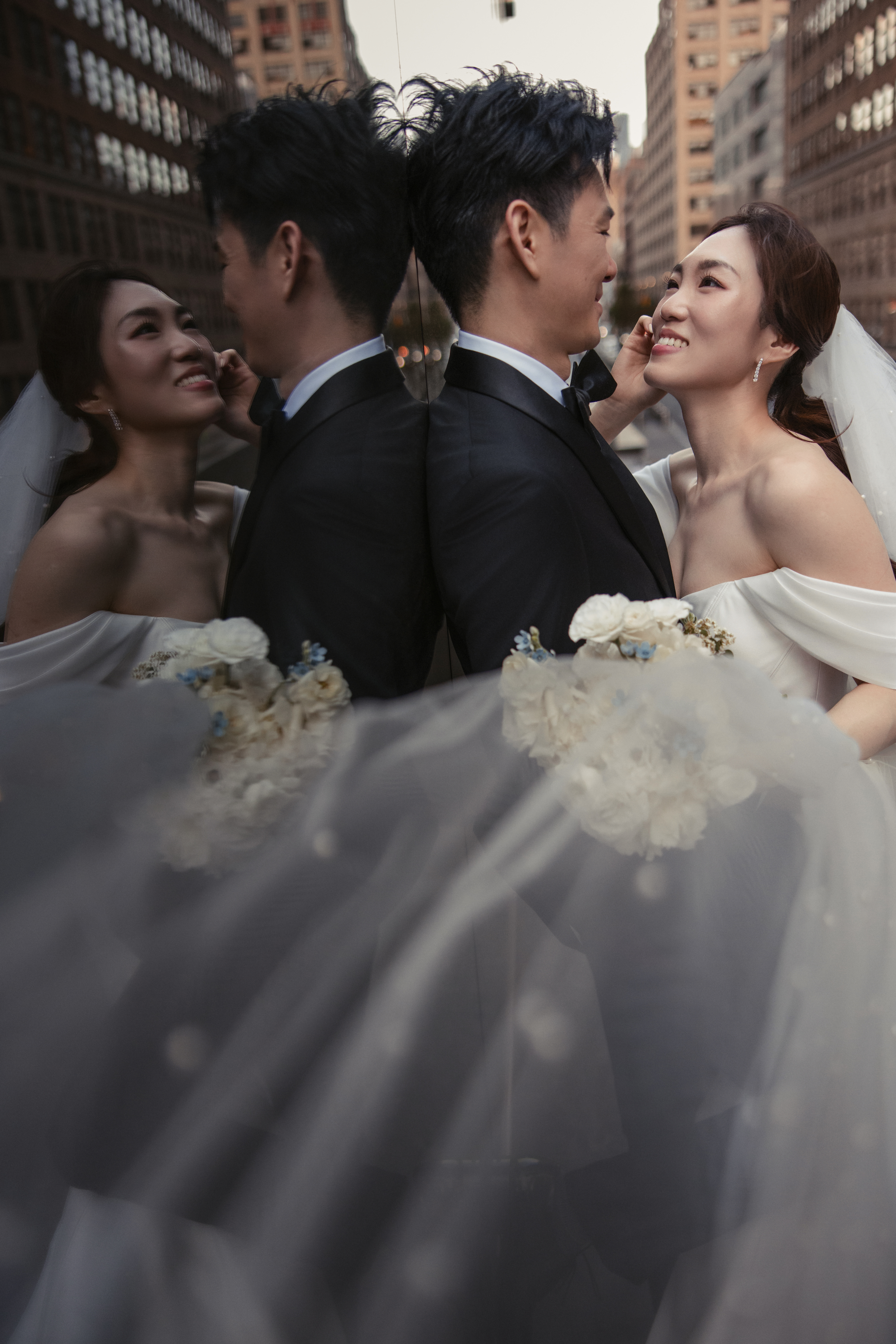 A bride and groom in wedding attire stand close together, smiling, with their reflection visible in a glass surface