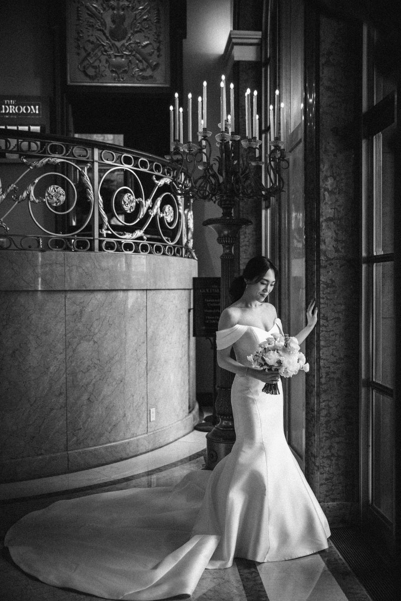 A bride in a long, off-the-shoulder gown stands by a large window holding a bouquet, with a grand staircase and candelabra in the background