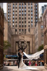 A bride and groom stand together at a wedding venue entrance, the bride's veil flowing gracefully, with St. Patrick’s Cathedral and a city skyline with tall buildings and onlookers in the background.