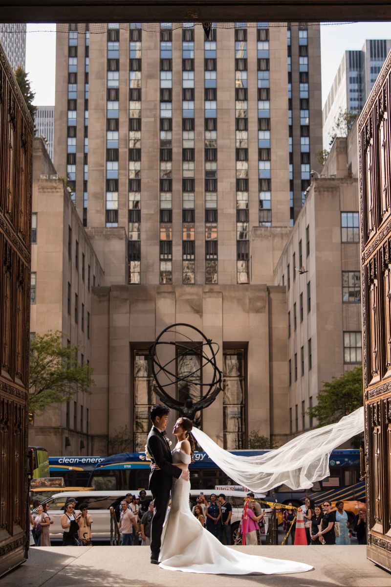 A bride and groom stand together at a wedding venue entrance, the bride's veil flowing gracefully, with St. Patrick’s Cathedral and a city skyline with tall buildings and onlookers in the background.
