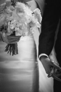 A bride holding a bouquet of flowers stands next to a man in a suit; they are holding hands at a wedding venue.