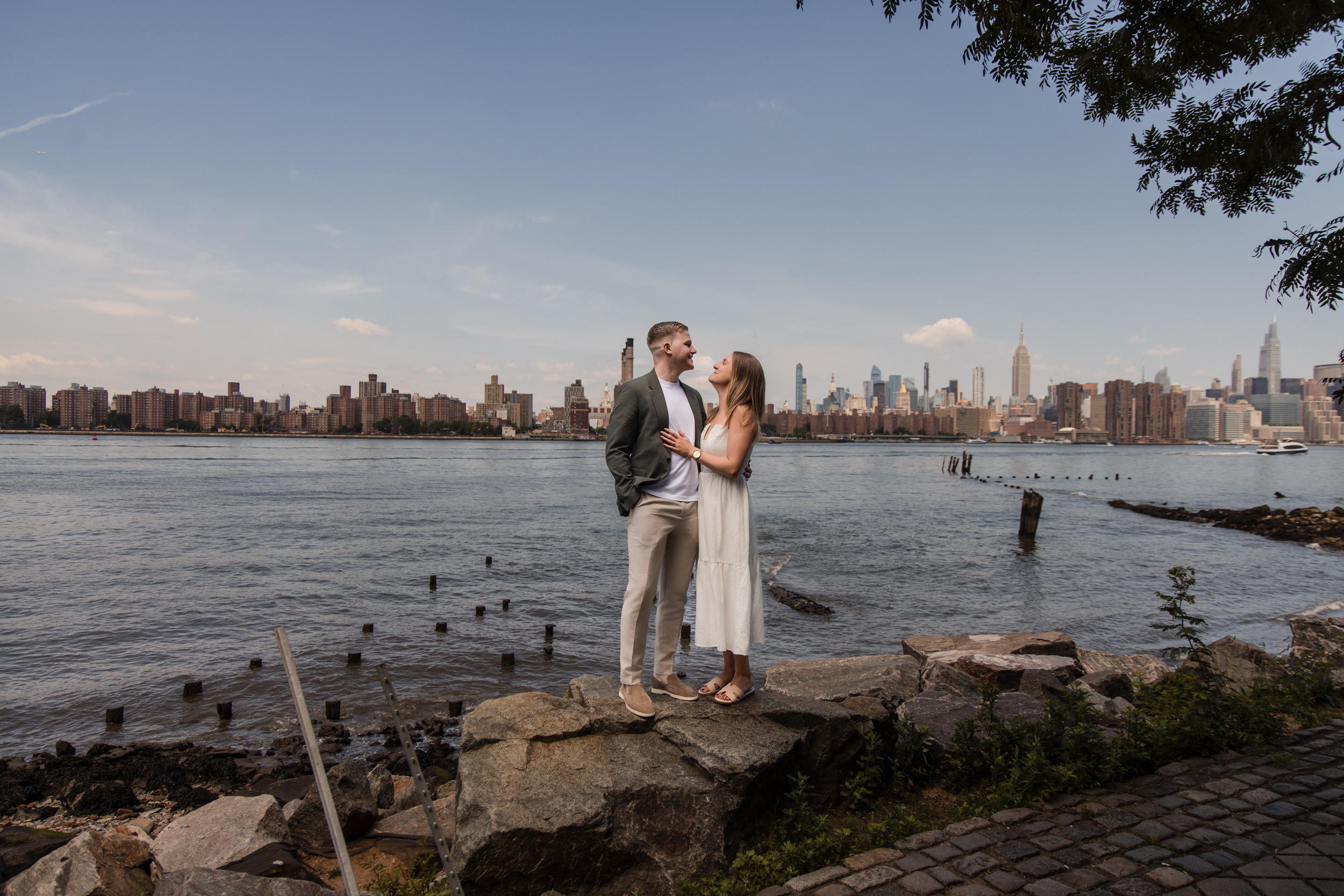 A couple stands on rocks by the water, facing each other, with a city skyline in the background under a partly cloudy sky—hire a NYC proposal photographer to capture moments like this.