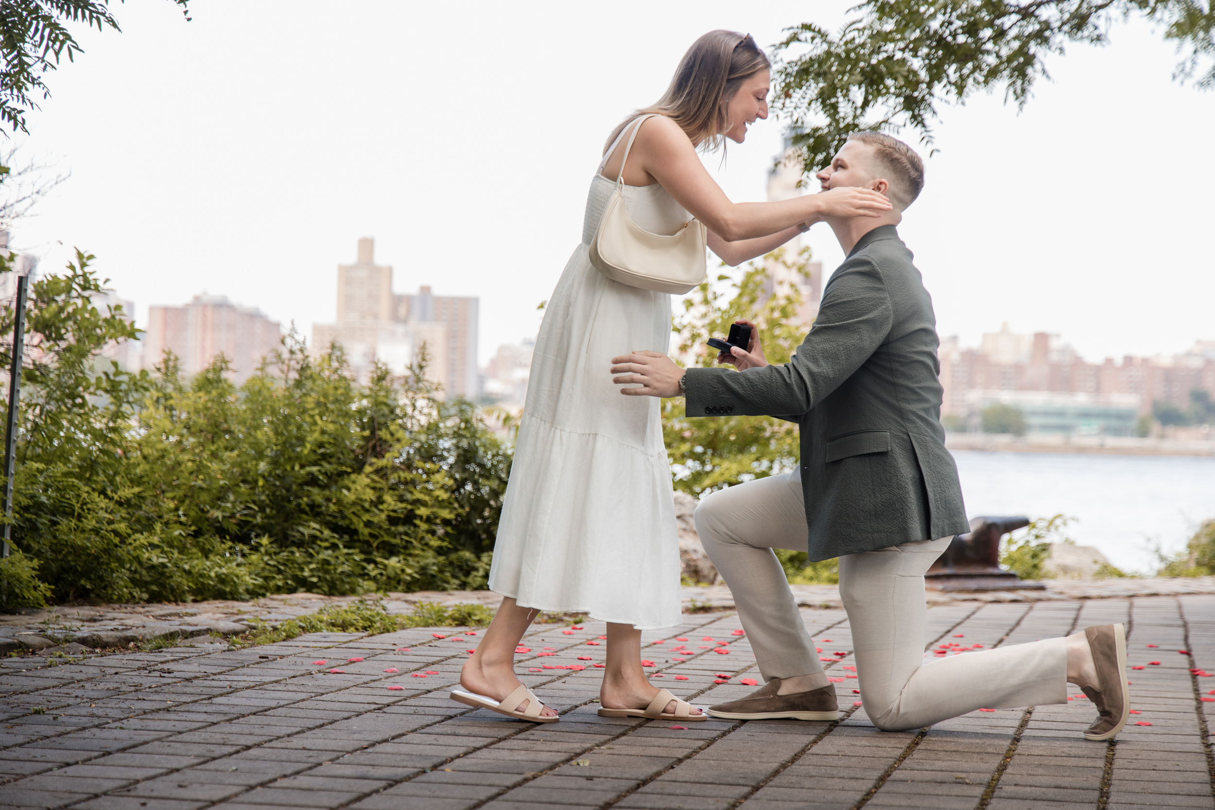 A man kneels and proposes to a woman outdoors near a river; she smiles, holding his face, with city buildings and greenery behind them—capturing the perfect moment for those wondering how to hire a proposal photographer in NYC.