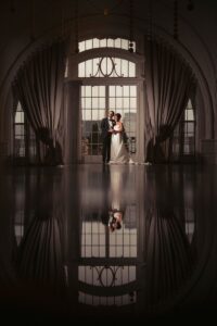 A bride and groom stand together in front of tall arched windows with curtains at, their reflection visible on the polished floor.