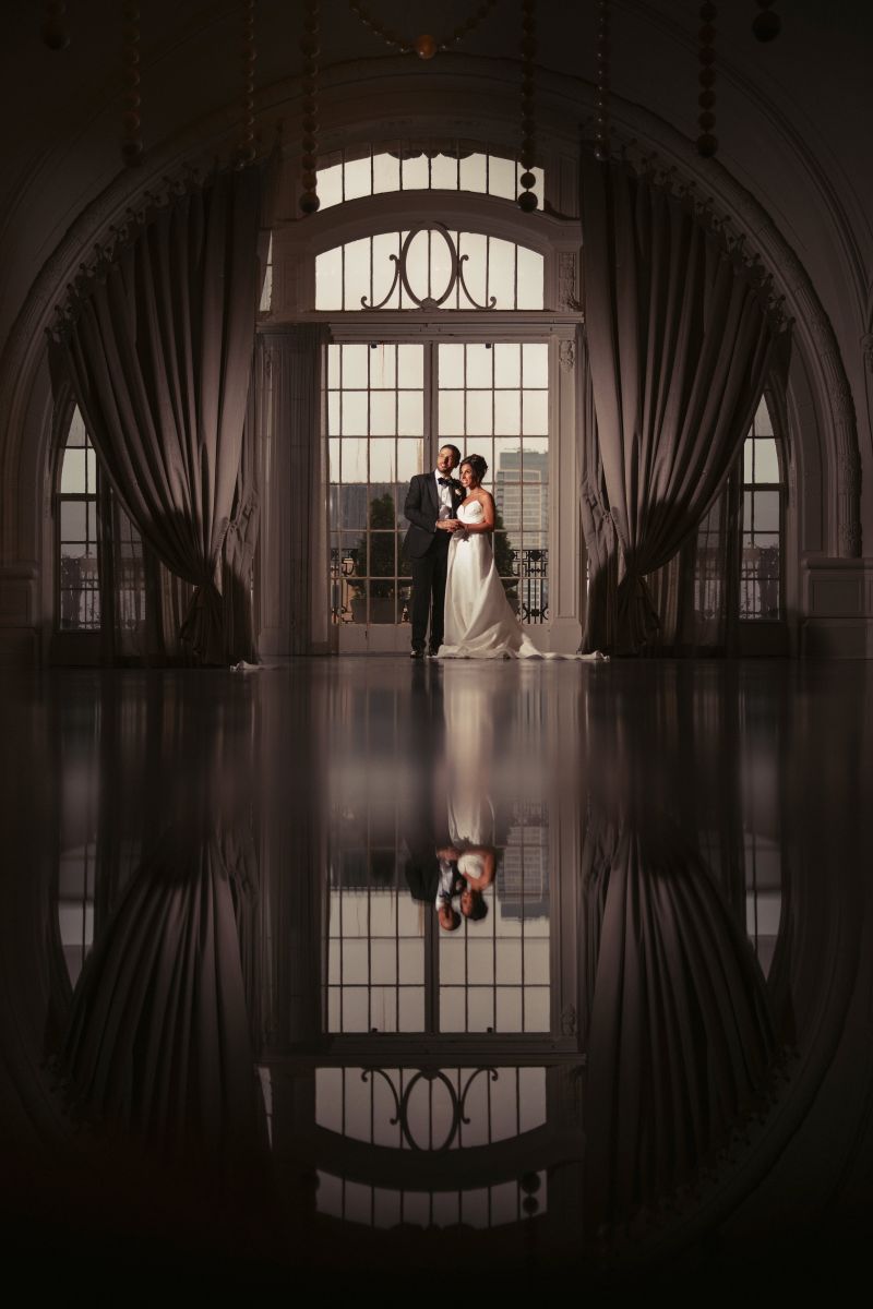 A bride and groom stand together in front of tall arched windows with curtains at, their reflection visible on the polished floor.