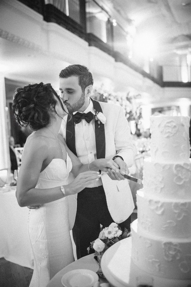 A bride and groom in formal attire cut a tiered wedding cake together while the groom kisses the bride on the cheek in a beautifully decorated indoor venue.