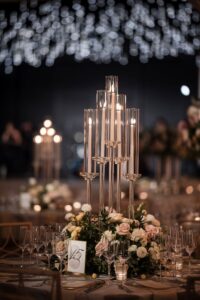 Elegant table setting at a wedding with tall glass candle holders, floral centerpiece, multiple wine glasses, and a table number card under soft, twinkling lights.