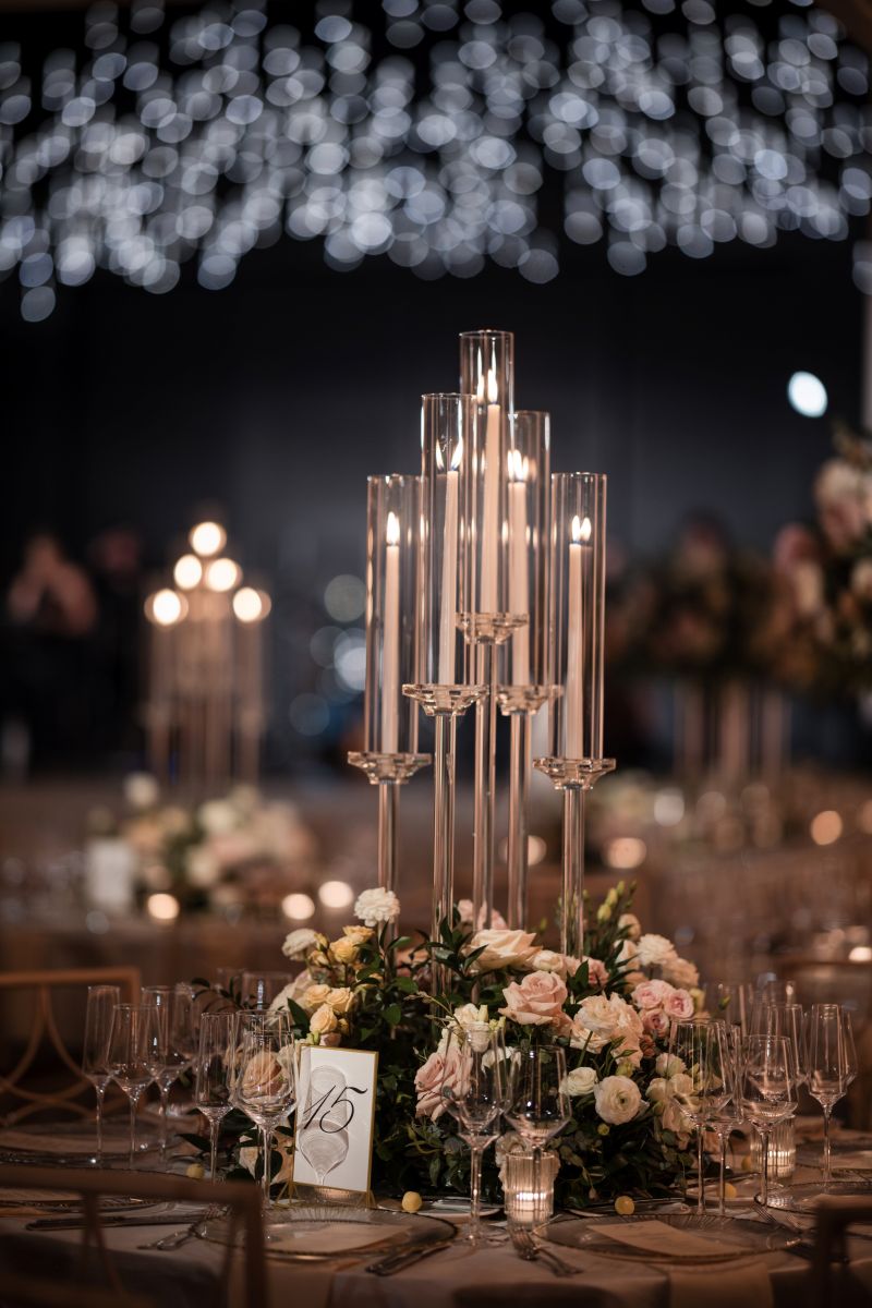 Elegant table setting at a wedding with tall glass candle holders, floral centerpiece, multiple wine glasses, and a table number card under soft, twinkling lights.