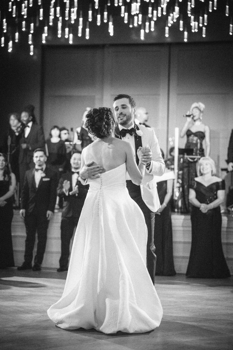 A bride and groom share a first dance at their wedding surrounded by elegantly dressed guests and a live band, under sparkling hanging lights.