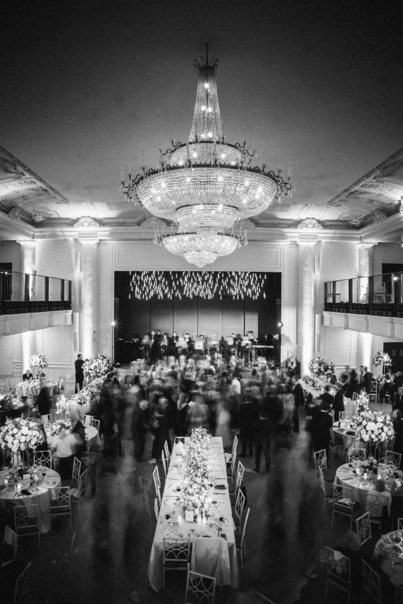 Black and white image of a wedding at The Grand Belle in Philadelphia with a large chandelier, decorated tables, and guests mingling near a stage with musicians.