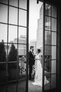 A bride and groom stand together on a balcony overlooking a cityscape, viewed through glass doors