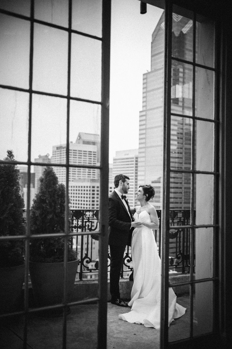 A bride and groom stand together on a balcony overlooking a cityscape, viewed through glass doors