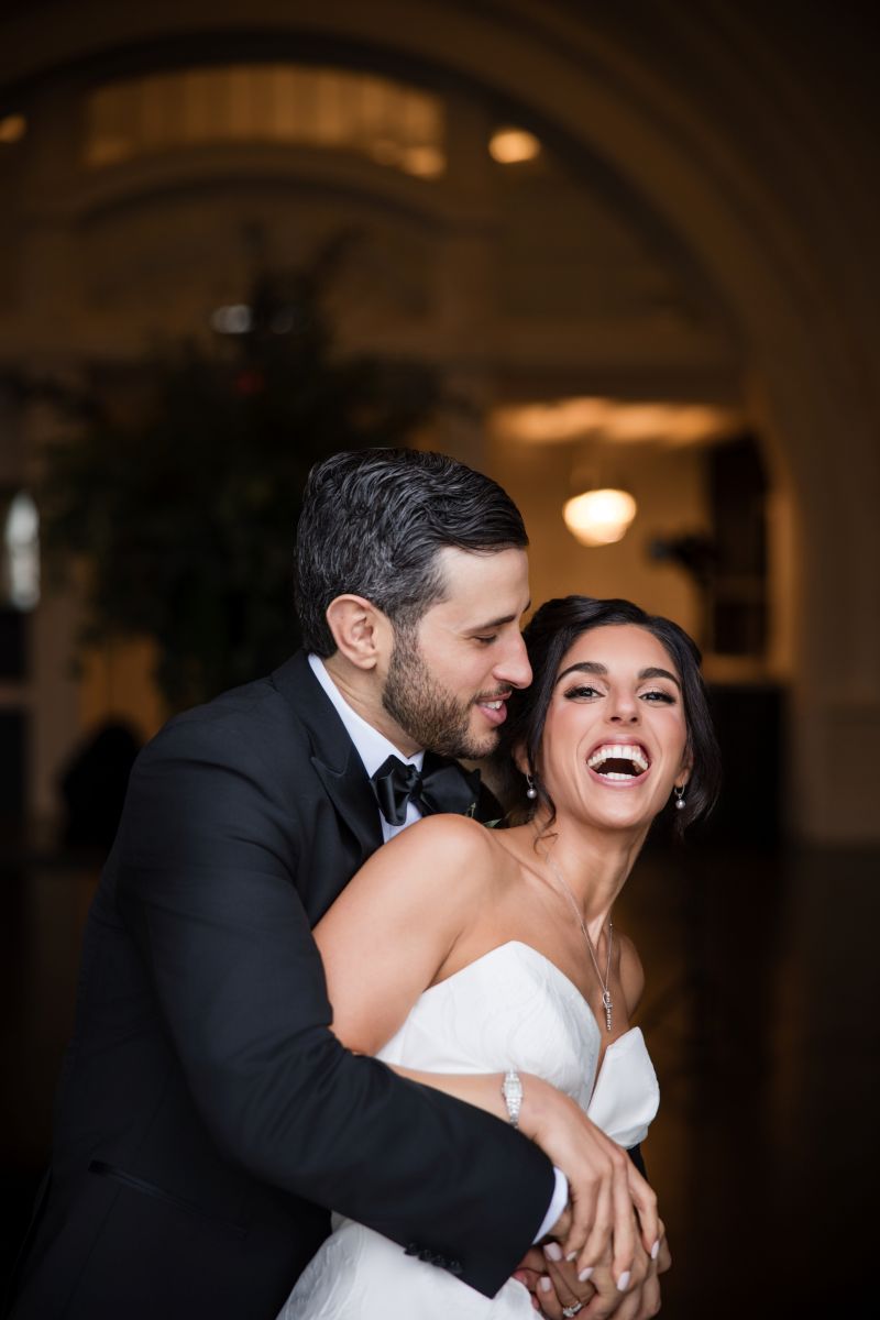 A man in a black tuxedo embraces a woman in a white wedding dress, both smiling and laughing in an elegant indoor setting