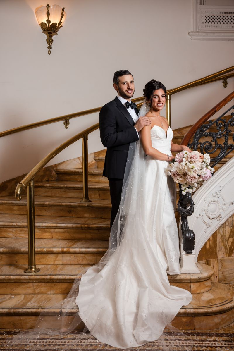 A bride in a white gown holding a bouquet stands beside a groom in a tuxedo on a marble staircase surrounded by ornate railings.
