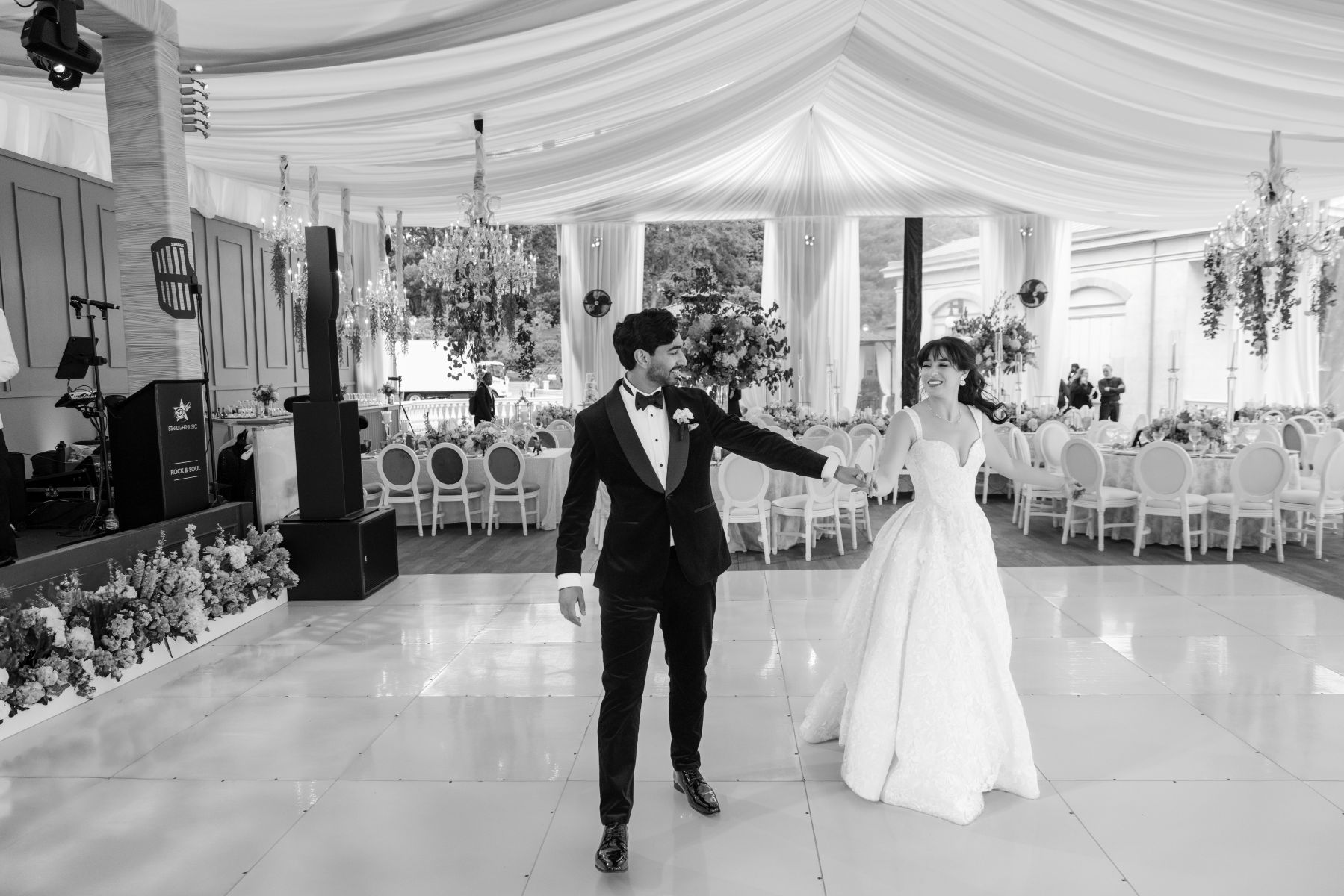 a couple on a dance floor holding hands and smiling at each other the woman is wearing a white wedding dress the man is wearing a black suit they are in a tent with tables, chairs, and chandeliers 