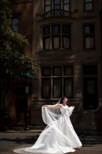 A bride in a white wedding dress stands in the street, holding her veil