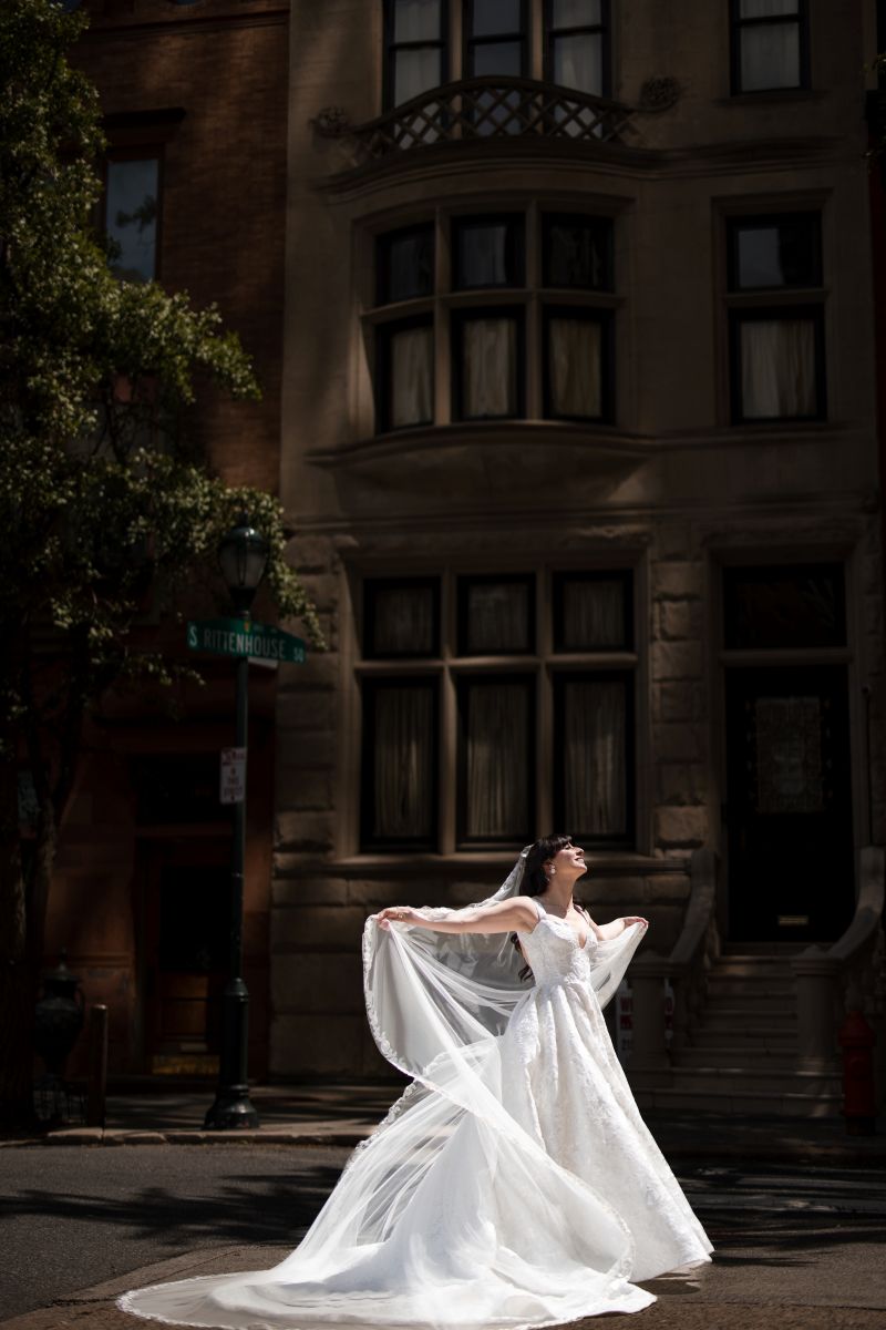 A bride in a white wedding dress stands in the street, holding her veil