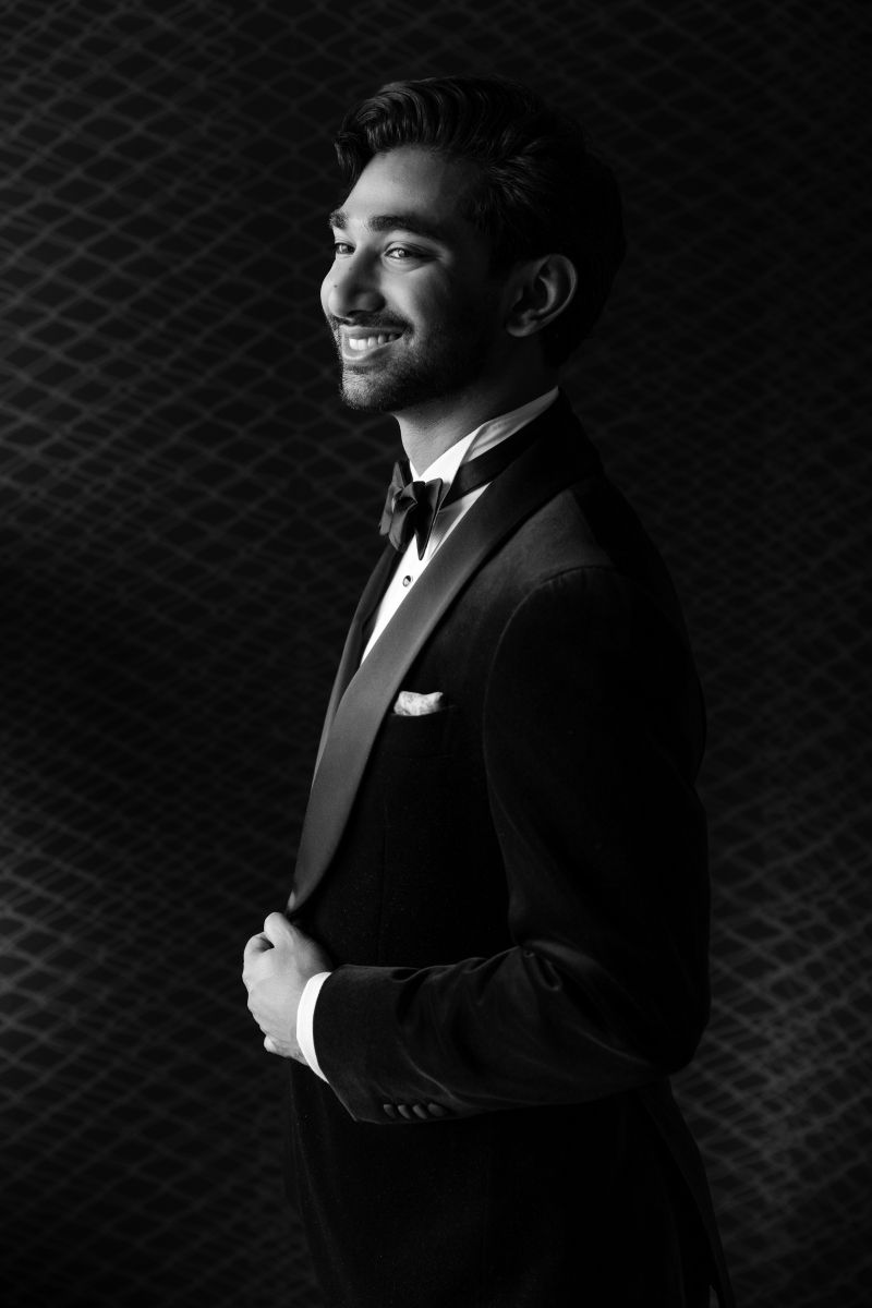 A man in a tuxedo and bow tie stands in profile, smiling, against a patterned background in a black and white photo