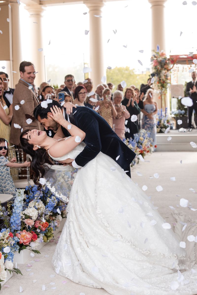 A bride and groom share a dip and kiss at their ceremony, surrounded by guests, flower arrangements, and falling white flower petals.