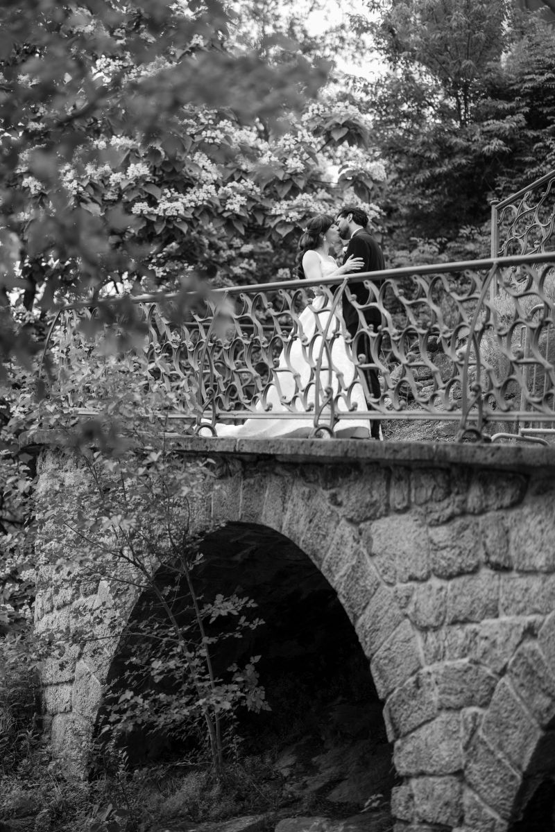A bride and groom share a kiss on a stone bridge surrounded by trees, captured in black and white